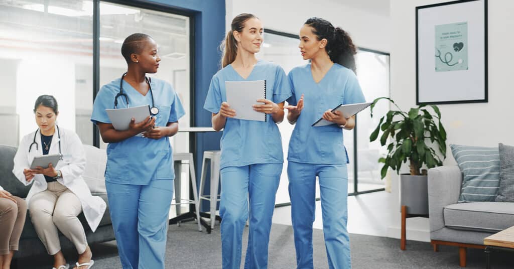 Three healthcare professionals in blue scrubs walk and converse in a medical facility. Another individual sits in the background, reading documents.