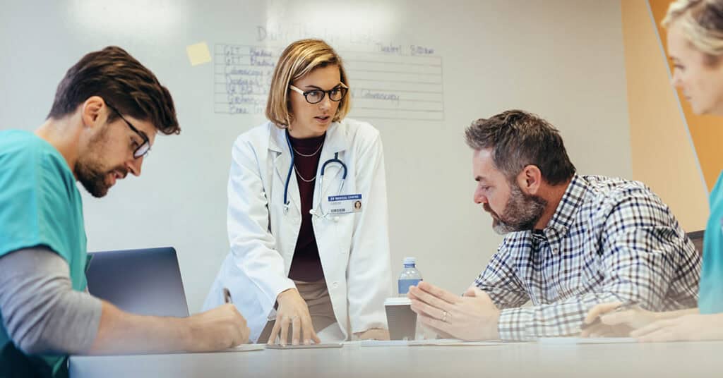 A doctor in a white coat talks to three colleagues seated around a table, discussing notes and papers.