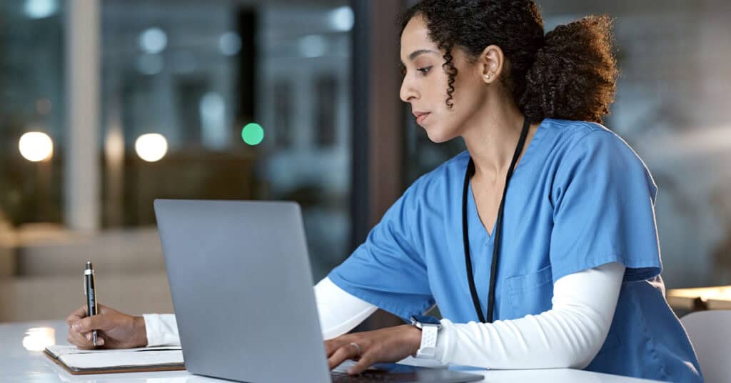 A person in blue scrubs is writing notes on paper while working on a laptop.