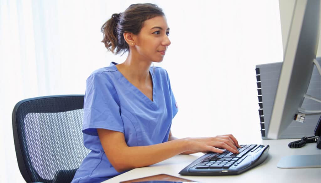 A woman in blue scrubs types on a keyboard while looking at a computer monitor.