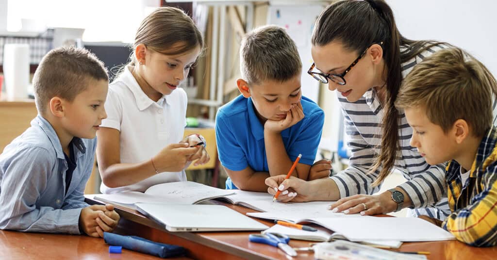 A teacher helps a group of four young students with their work at a table in a classroom.