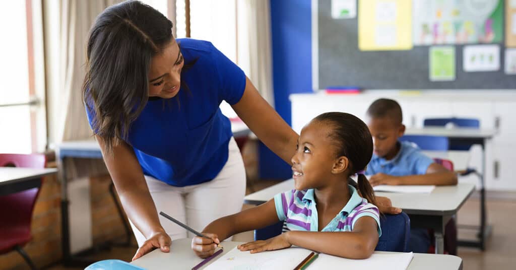 A woman in a blue shirt is helping a child with schoolwork in a classroom. Another child is sitting in the background.