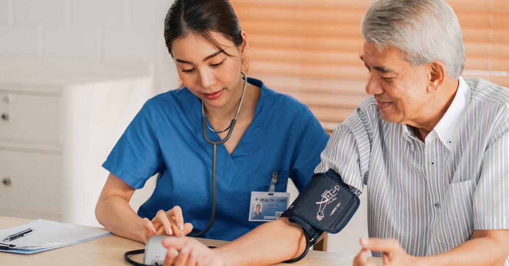 A healthcare professional in blue scrubs checks the blood pressure of an older man using a digital blood pressure monitor.