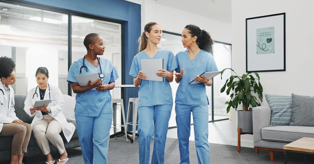 Three healthcare professionals in blue scrubs walk and discuss in a medical office, while two other staff members are seated and working on tablets in the background.