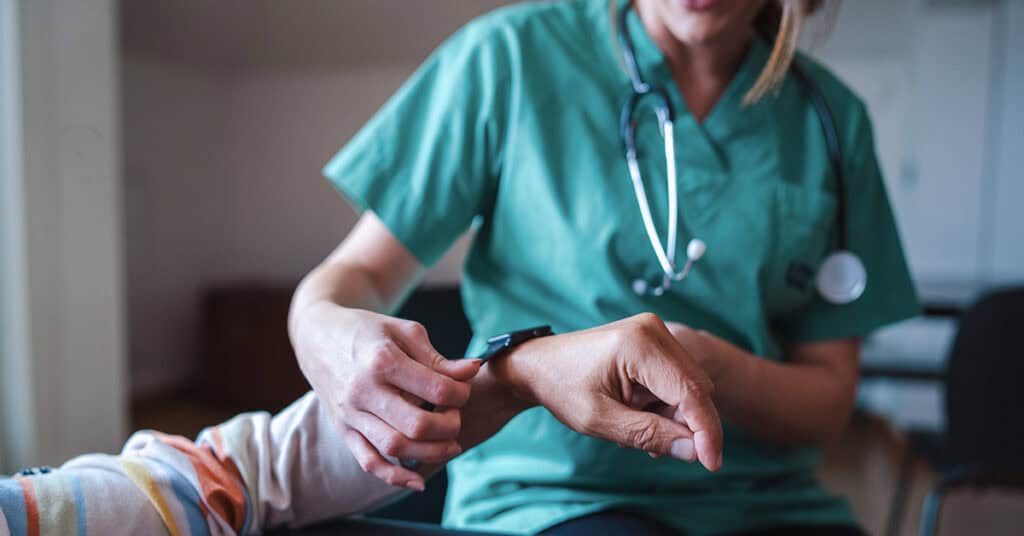 A healthcare professional in a green uniform measures a patient's pulse using a pulse oximeter on the patient's wrist.
