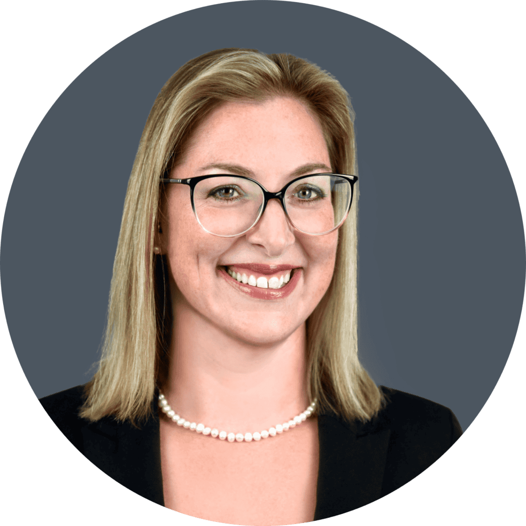 A woman with shoulder-length blonde hair and glasses is smiling. She is wearing a black blazer and a pearl necklace, against a gray background.
