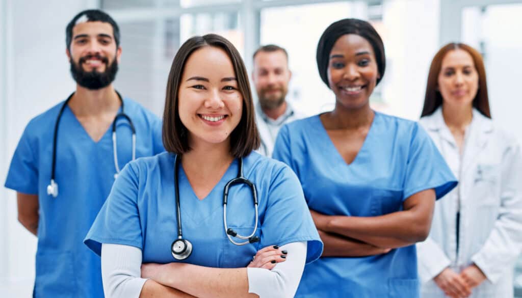 A group of five healthcare professionals in blue uniforms and white lab coats stand together, smiling confidently, with stethoscopes visible on some of them.