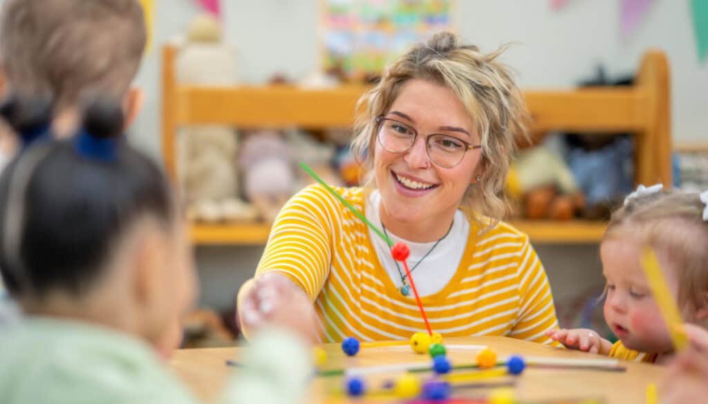 A woman wearing glasses and a yellow striped shirt interacts with children at a table covered with craft materials in a classroom.