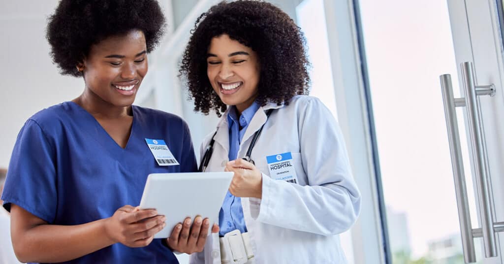 Two healthcare professionals, one in blue scrubs and the other in a white lab coat, smiling and looking at a tablet in a hospital setting.