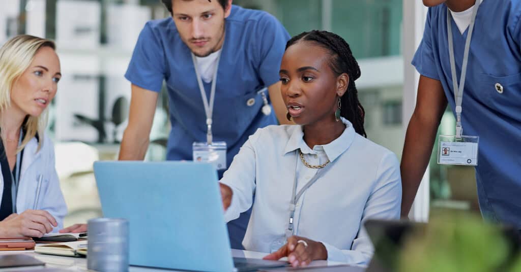 A woman in a white shirt points at a laptop screen on a table, surrounded by three people in blue scrubs, during a discussion in a well-lit room.