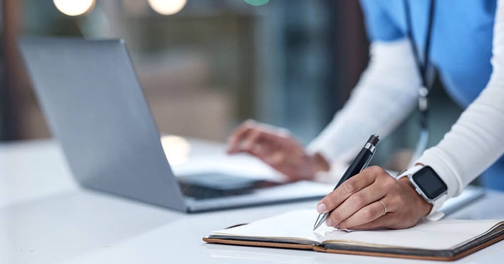 Person wearing a smartwatch and a blue shirt, writing in a notebook with a pen, while using a laptop on a white desk.