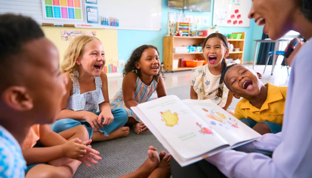 A group of children sit on the floor, smiling and laughing, while an adult reads a picture book to them in a brightly lit classroom.