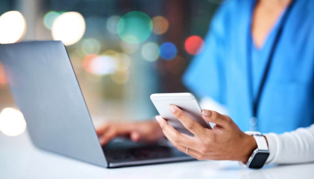 A person in medical scrubs uses a smartphone while working on a laptop, with a background of blurry lights.
