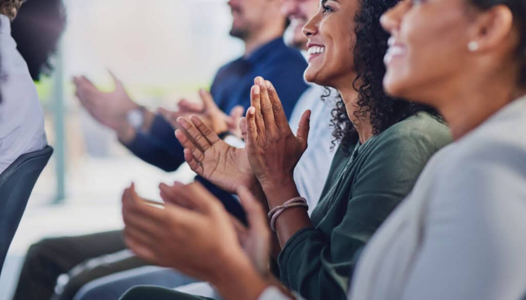 A group of people sitting in chairs are clapping and smiling, appearing to be engaged in a positive event or presentation.