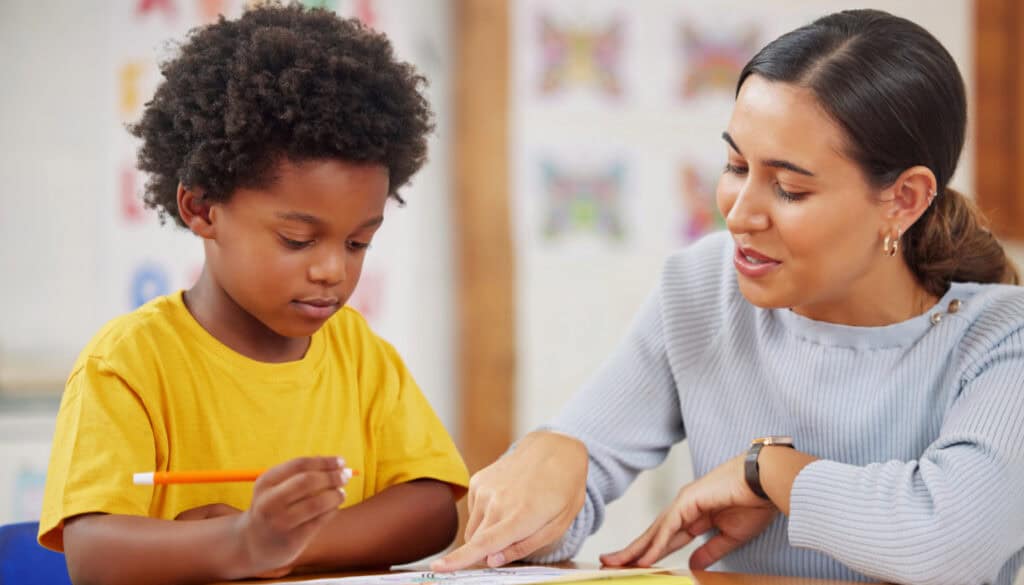 A child in a yellow shirt and a woman with brown hair are sitting at a table while engaging in a drawing activity. The woman is guiding the child as they both focus on the drawing.