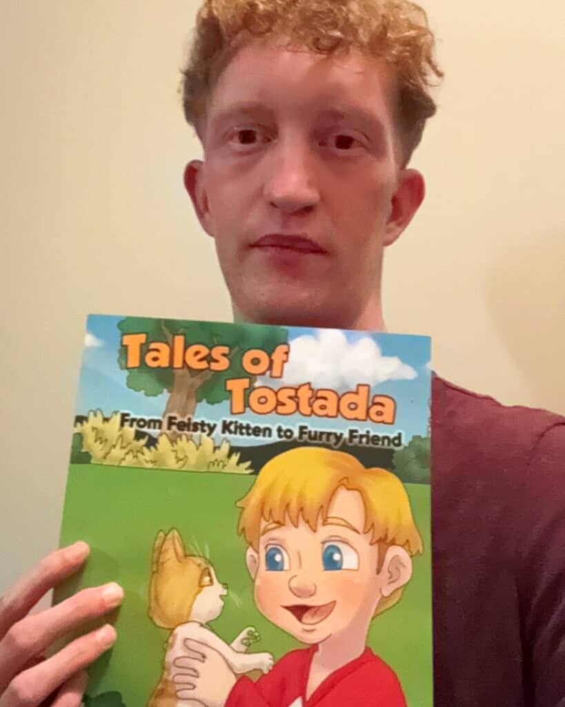 Person holding a children's book titled "Tales of Tostada: From Feisty Kitten to Furry Friend" with a cover illustration of a child and a kitten.