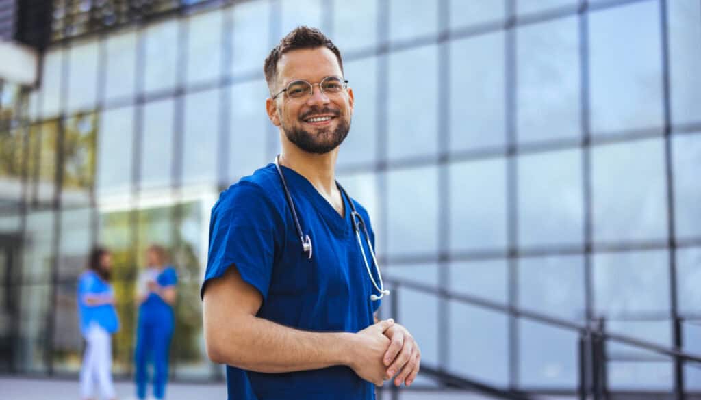 Medical professional working in a hospital. He is dressed in scrubs looking at the camera smiling with a stethoscope around his neck. Happy in his Profession. Male nurse with stethoscope standing at clinic.