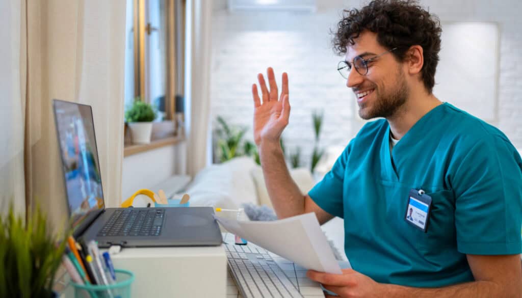 A man in medical scrubs waves at a laptop screen during a video call in a bright room with plants. He holds papers in his other hand.