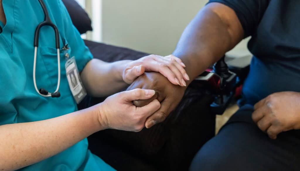 A healthcare worker in a teal uniform holds the hand of a person in a black shirt, providing reassurance or support.