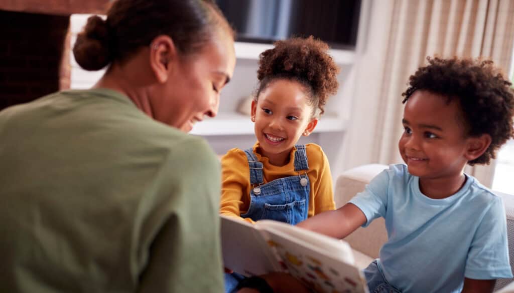A woman reads a book to two smiling children sitting on a couch.