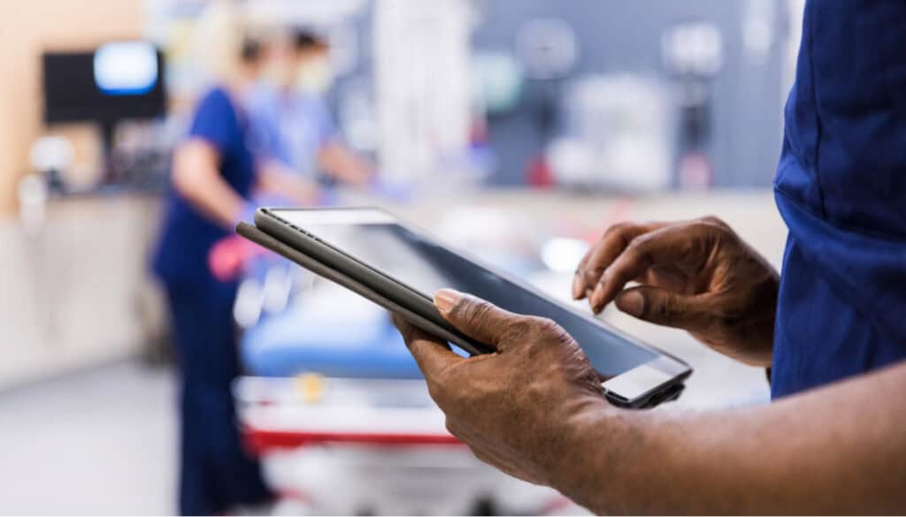 Person in medical scrubs using a tablet in a healthcare setting, with blurred figures in the background attending to a patient on a bed.