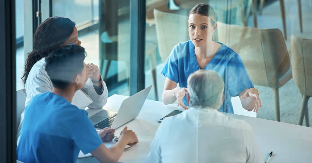 A group of healthcare professionals in blue scrubs and a white coat are having a discussion around a table with a laptop.