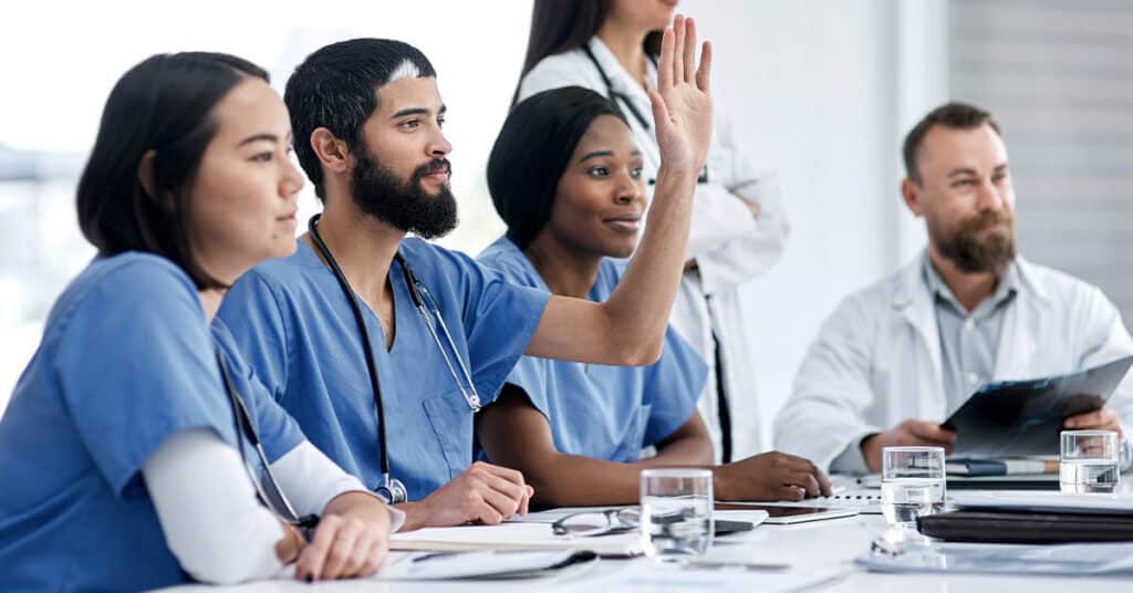 Medical professionals in a meeting, with one person raising a hand. They are sitting at a table with papers and glasses of water.