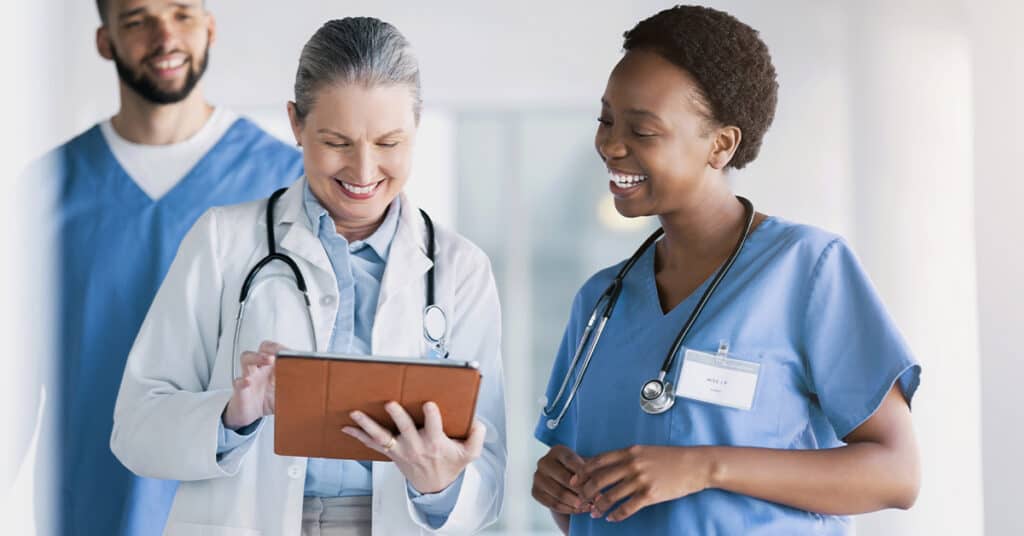 Three healthcare professionals, two women and one man, smiling and engaged in a discussion. One woman is holding a tablet, and all are wearing medical uniforms with stethoscopes.
