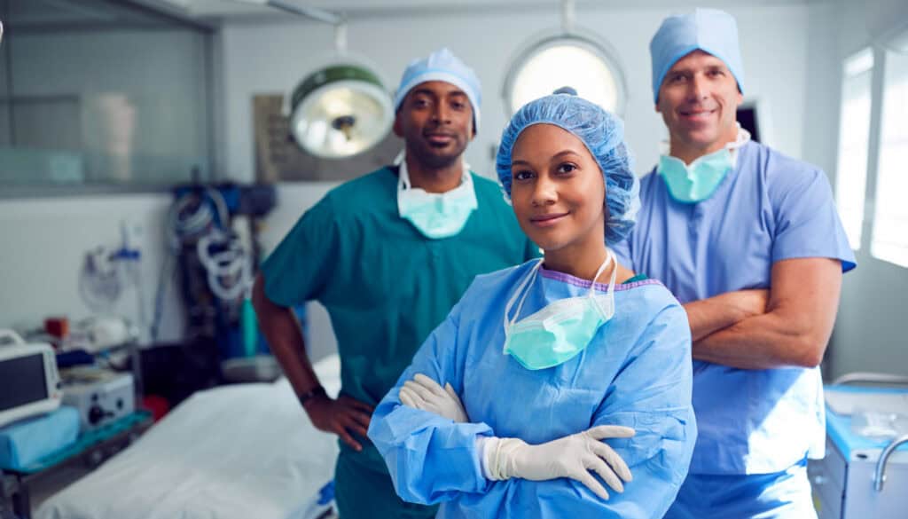 Three medical professionals in surgical scrubs pose confidently in an operating room setting, with medical equipment and a surgical table in the background.