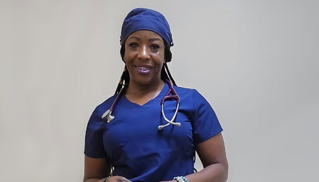 A person wearing blue medical scrubs and a matching cap, with a stethoscope around their neck, smiles at the camera.