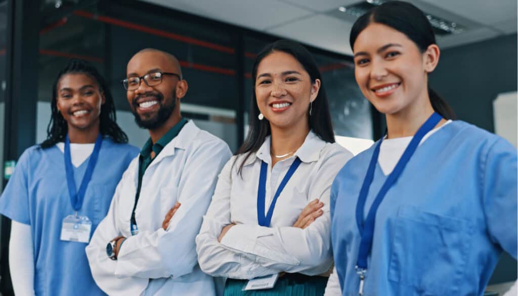 Four healthcare professionals, two in medical scrubs and two in business attire, stand smiling with arms crossed in a hospital setting.