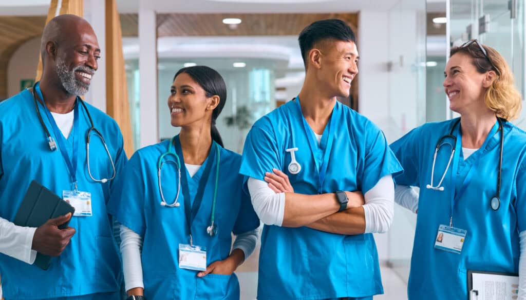 A diverse group of four medical professionals in blue scrubs are smiling and conversing in a hospital hallway.