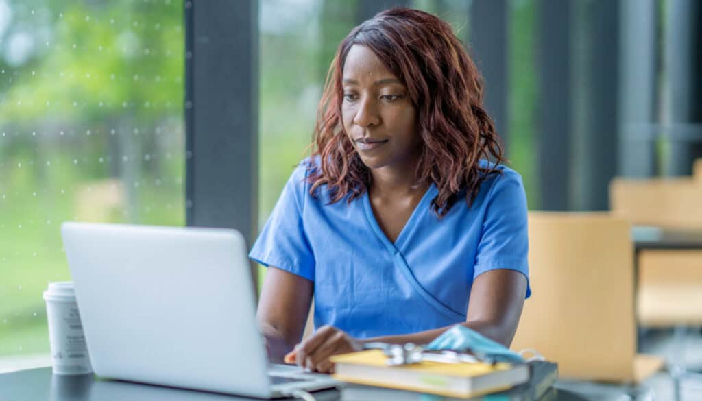 A healthcare professional in blue scrubs works on a laptop in a bright room, with a coffee cup and notebooks on the table.