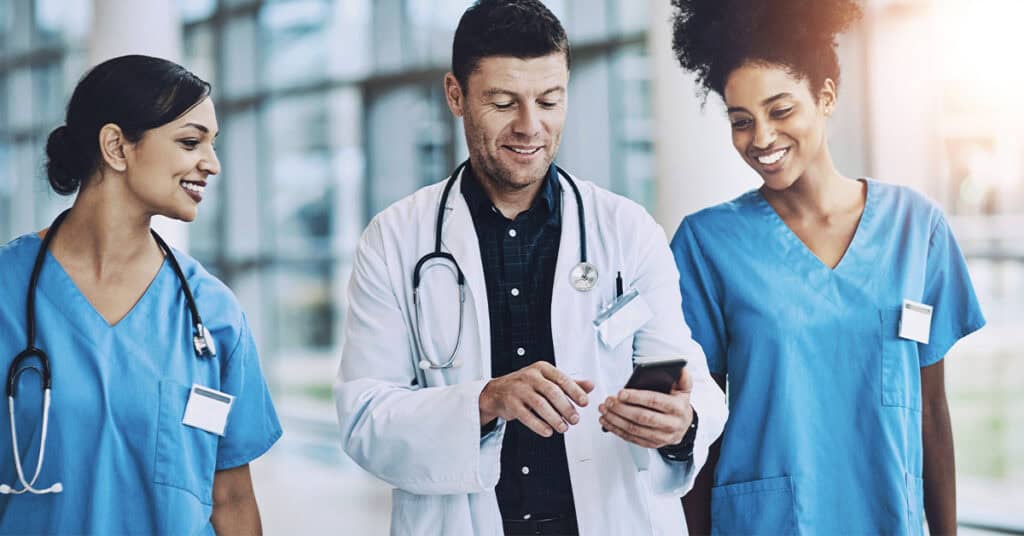A doctor and two nurses in scrubs are walking in a hospital corridor. The doctor is looking at a smartphone and smiling, and the nurses are also smiling.