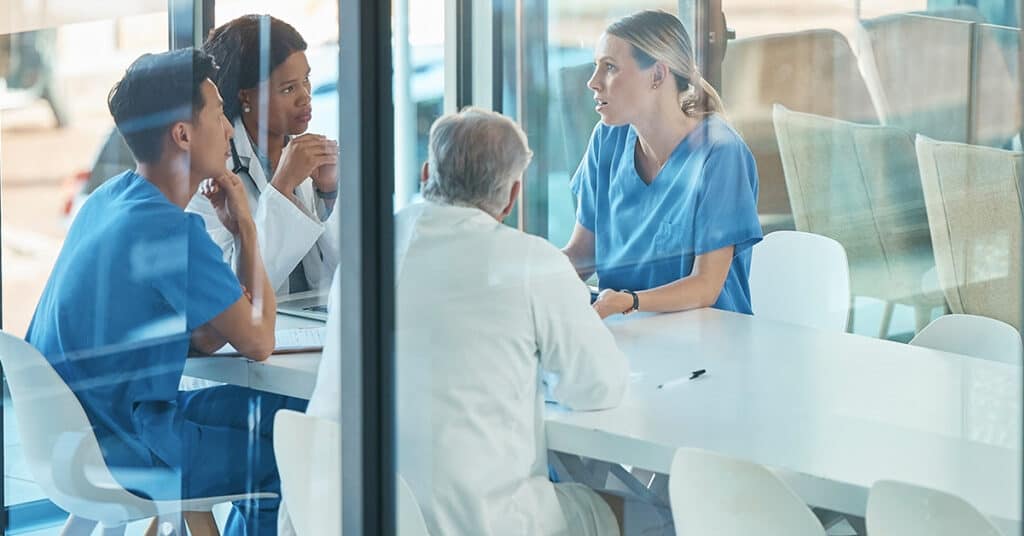 Four medical professionals in scrubs and white coats are seated around a table in a glass-walled meeting room, engaged in a discussion.