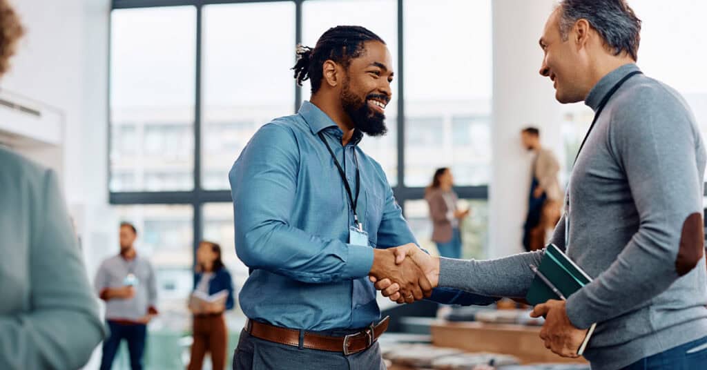 Two men are shaking hands and smiling in an office setting, with people conversing in the background.