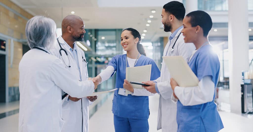 Medical professionals in a hospital hallway, engaging in conversation. A doctor and a nurse shake hands, while two other nurses hold documents and stand nearby, smiling.