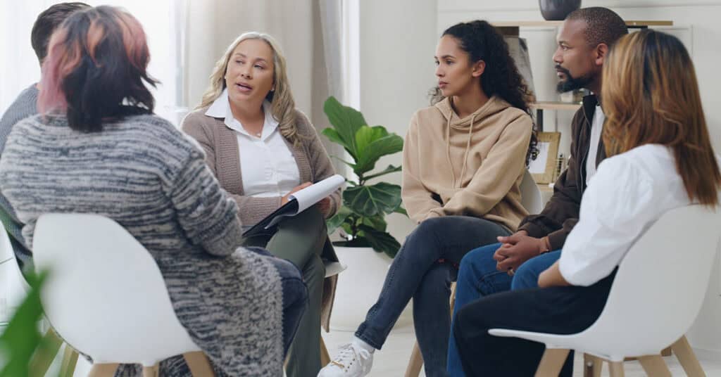 A group of people sit in a circle, engaged in a discussion. Some hold notebooks and wear casual attire. A plant and bookshelves are in the background.