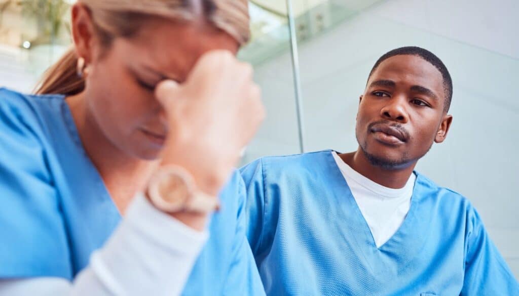 Two healthcare professionals in blue uniforms. One person looks down, appearing stressed, with a hand on their forehead. The other person sits beside, watching attentively.