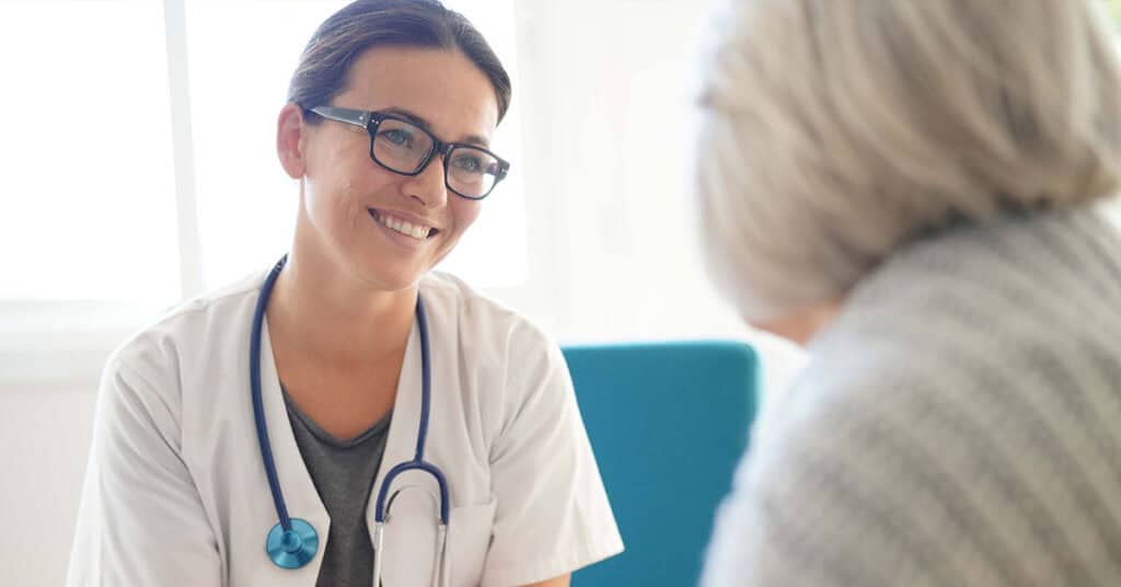 A doctor wearing glasses and a stethoscope smiles while talking to an older patient in an office setting.