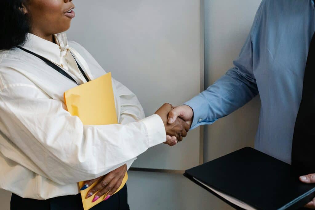 Two people in business attire are shaking hands. One person holds a yellow folder, while the other holds a black folder.