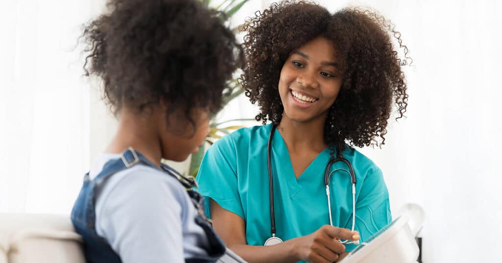 A healthcare worker in scrubs with a stethoscope talks to a child sitting on a sofa, holding a clipboard.