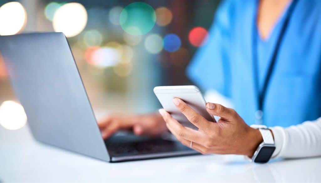 Person in blue scrubs using a smartphone and laptop at a desk.