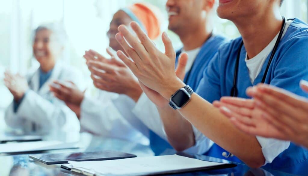 Healthcare professionals in scrubs and lab coats sit at a table, clapping. A tablet and notebook are visible in the foreground.