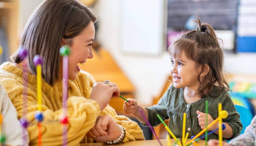 A woman and a young girl sit at a table, smiling at each other, surrounded by colorful educational toys.