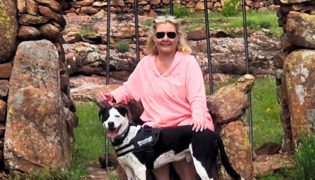 A woman in sunglasses and a pink shirt sits on a stone structure with a black and white dog wearing a harness in front of her.