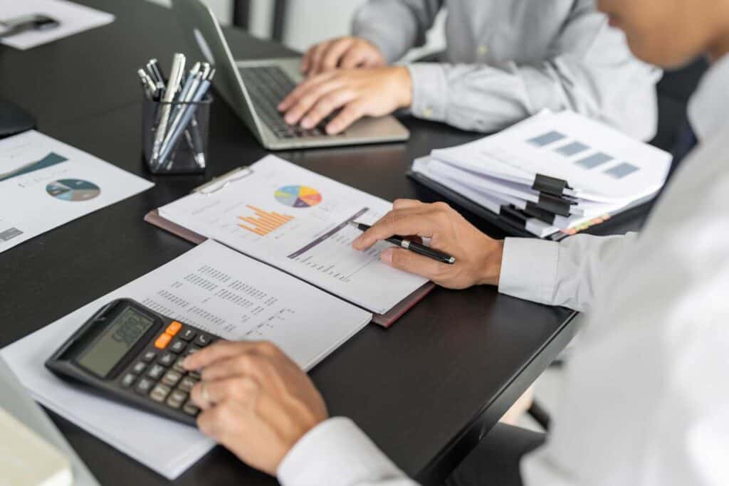 Two people working at a desk with laptops and documents. One uses a calculator; the other reviews a chart. Papers and office supplies are scattered on the dark tabletop.