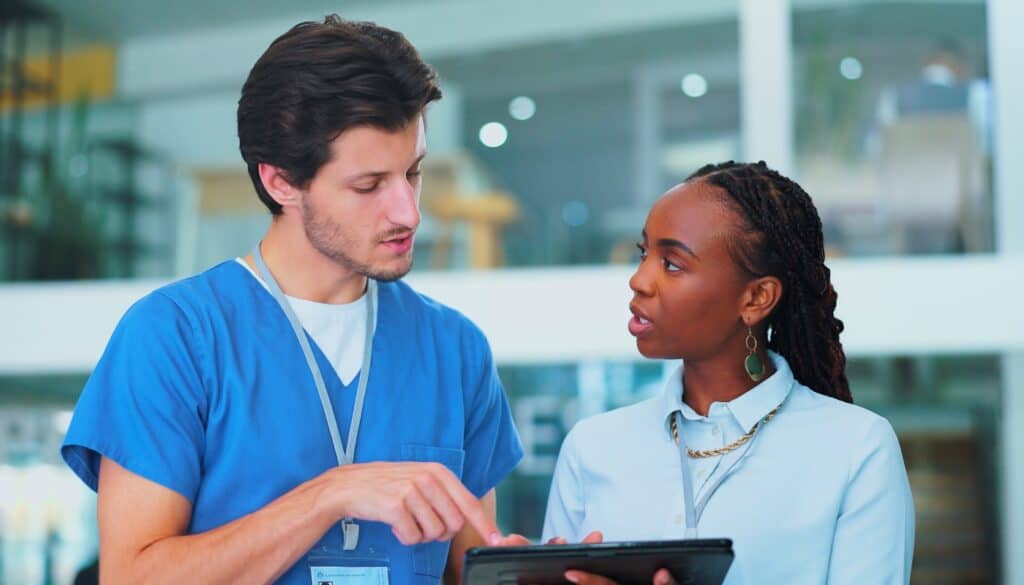 A man in blue scrubs discusses something on a tablet with a woman in a light blue shirt. They are in a brightly lit indoor setting.