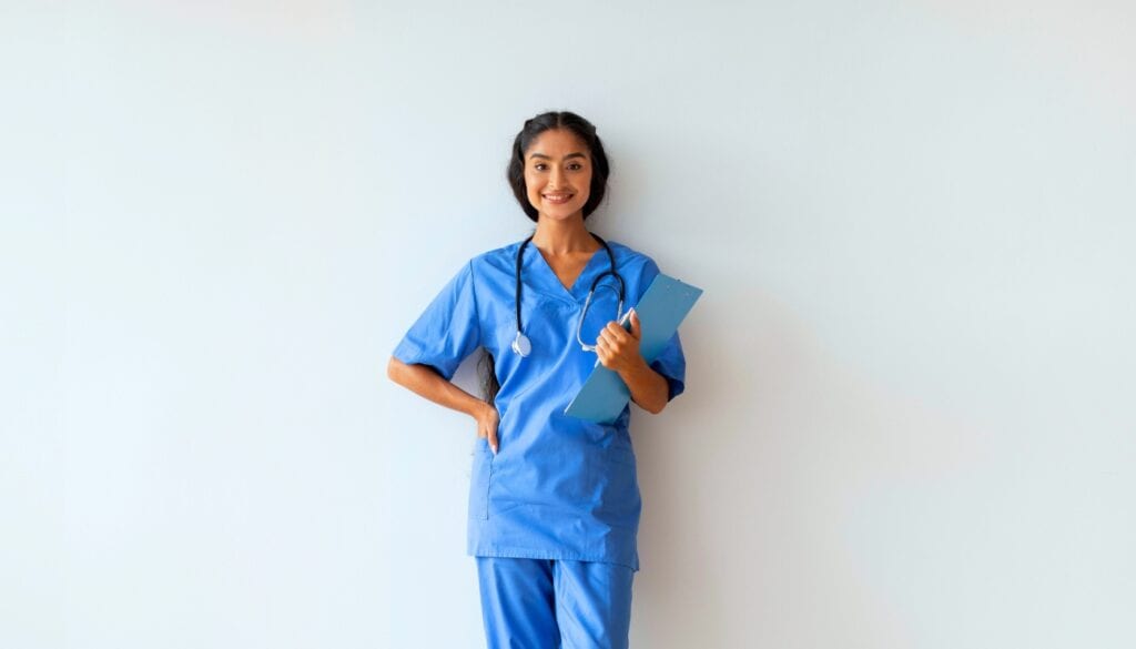 A person in blue scrubs, holding a clipboard and stethoscope, smiles while standing against a plain background.