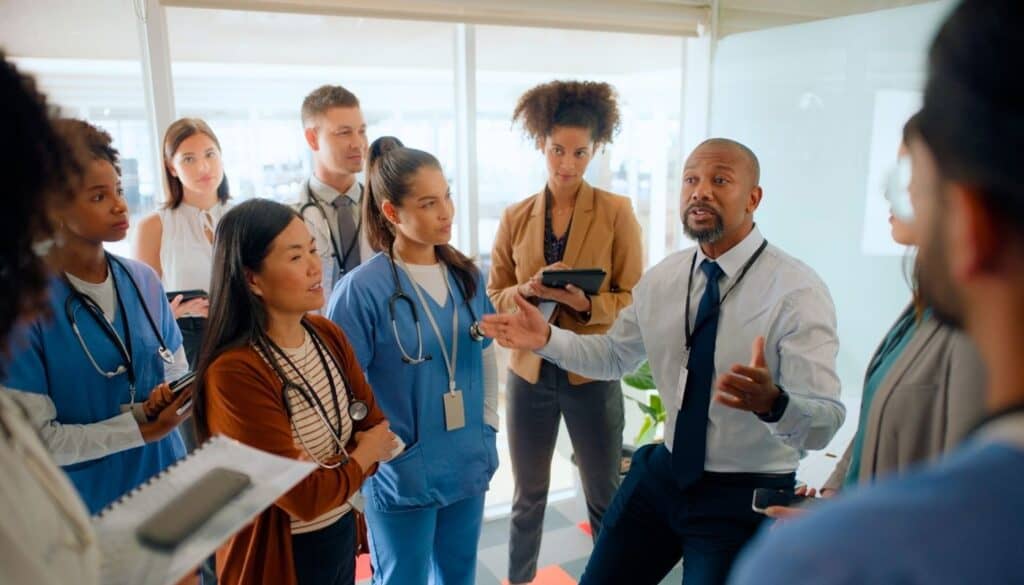 A diverse group of medical professionals and office workers engaging in a discussion in a bright room. Some hold notepads or tablets. A man in a tie is speaking, and others attentively listen.